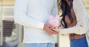 Couple holding a piggy bank in their new home. Home savings concept. They are standing in front of the their new modern house. Both are happy and smiling.